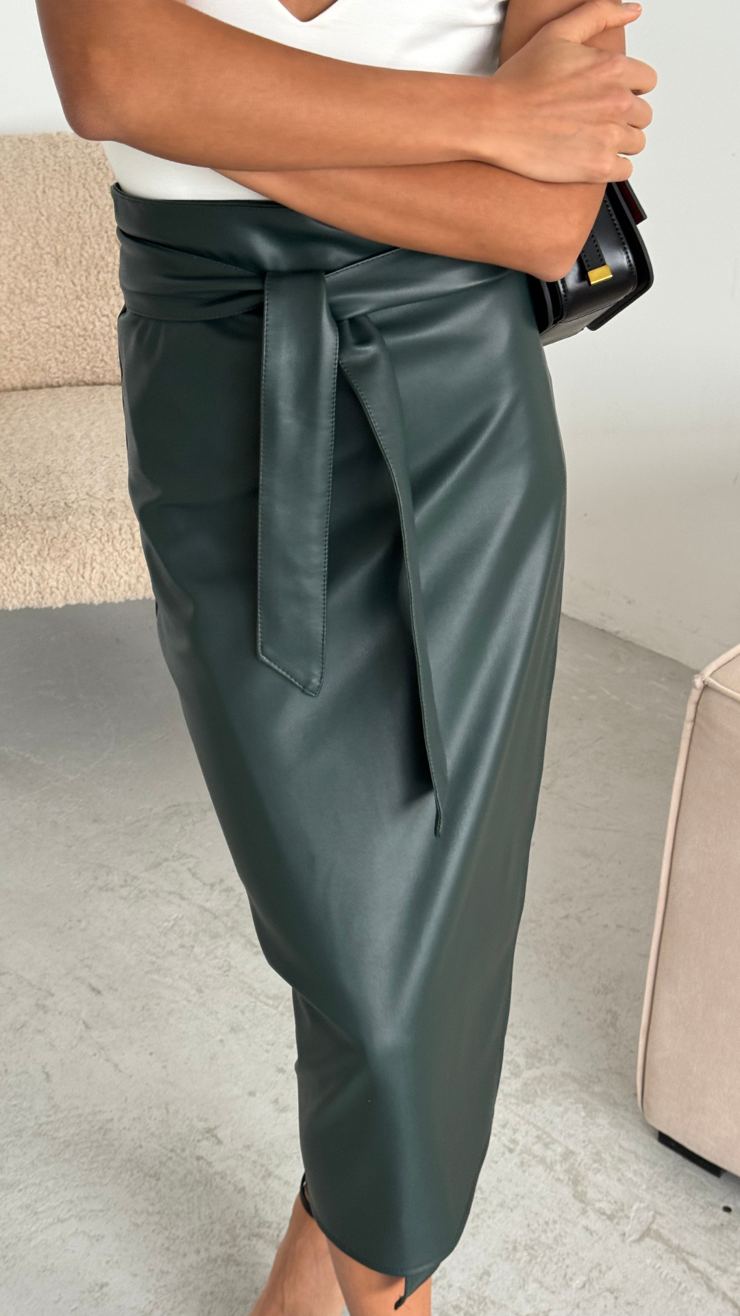 dark green faux leather midi skirt with a wrap design and tie waist, styled with a white top and accessorized with a black handbag