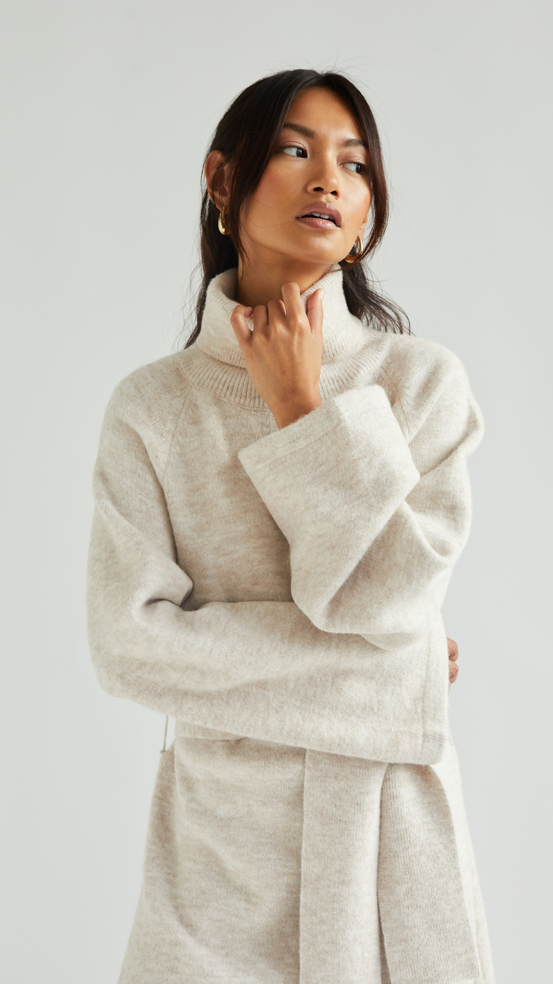 beige turtleneck sweater dress with a tie-waist detail and wide sleeves, accessorized with gold hoop earrings