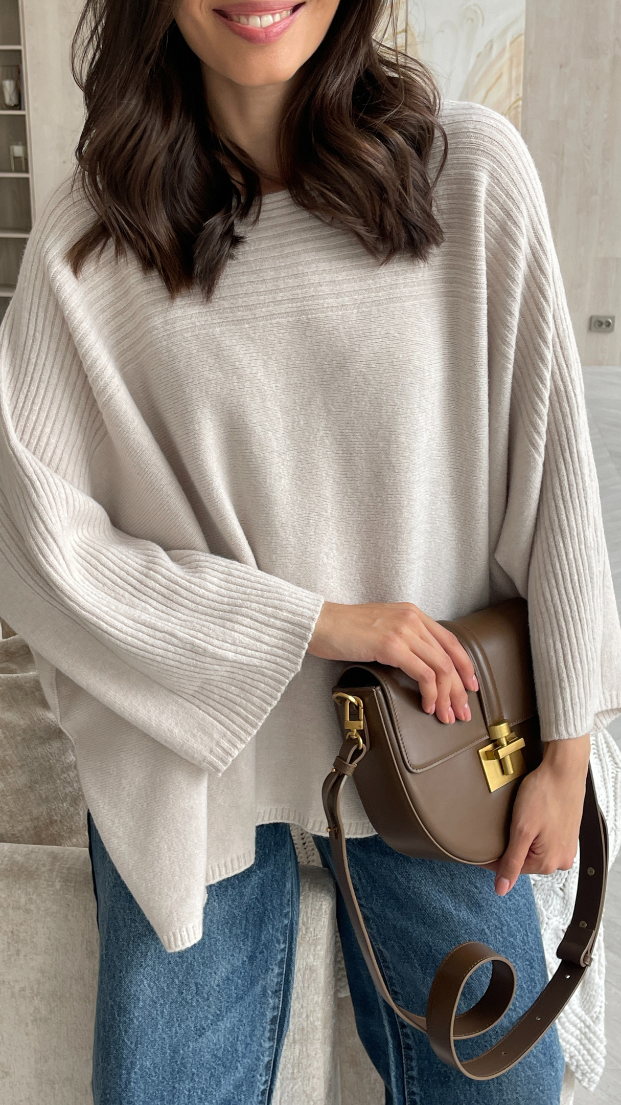 Woman wearing an oversized beige poncho sweater with ribbed detailing on the sleeves, paired with blue jeans and accessorized with a brown leather handbag featuring a gold clasp.