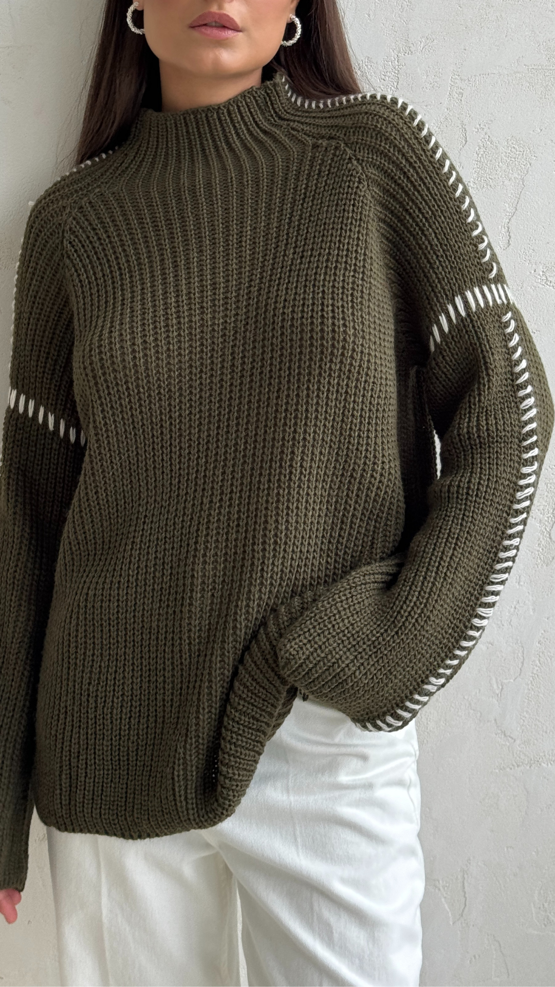 Woman wearing an olive green chunky knit sweater with white stitched detailing along the shoulders and sleeves, paired with white trousers
