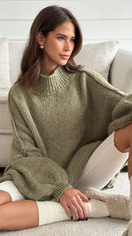 Woman sitting on a plush beige sofa, wearing an oversized olive green knit sweater with voluminous sleeves, paired with white ribbed shorts and cozy cream-colored socks