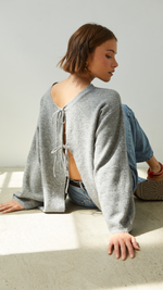 Woman sitting on the floor wearing a grey open-back sweater with delicate tie details, paired with casual blue jeans and sneakers