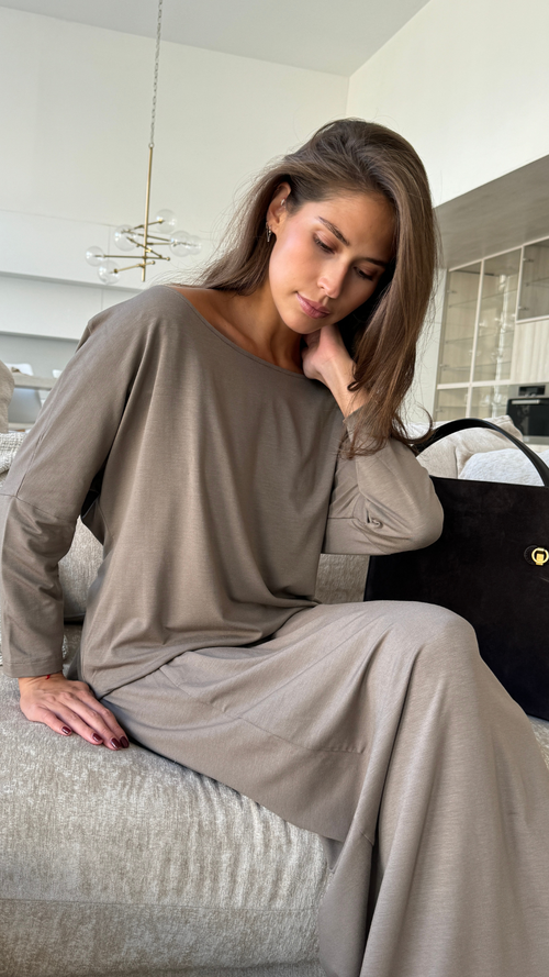 taupe long-sleeve top and long skirt, seated on a sofa in a modern living space, with a black handbag