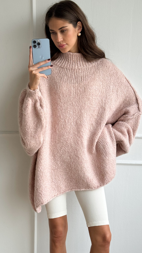 Woman wearing an oversized blush pink knit sweater with a high neckline and balloon sleeves, paired with white ribbed leggings