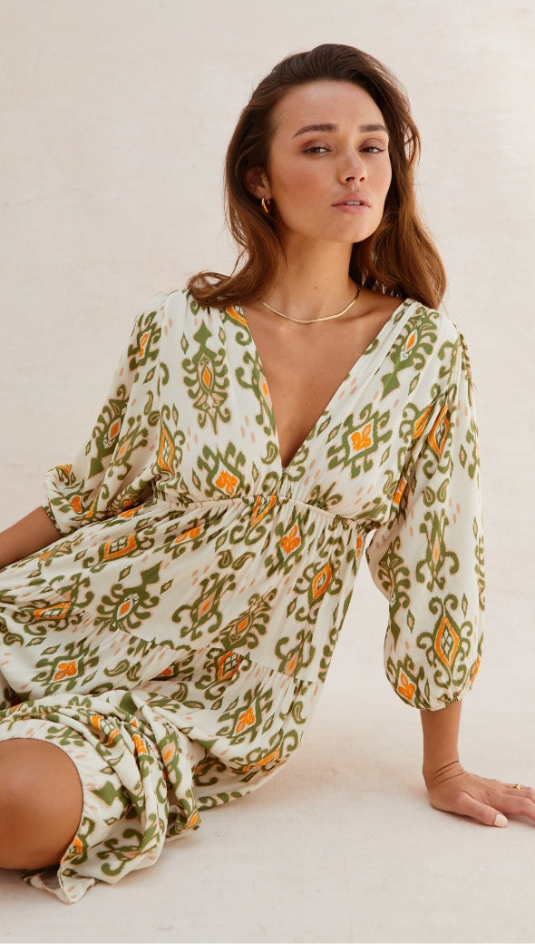 cream midi dress with a green and orange ikat-inspired pattern, featuring a deep V-neckline, three-quarter puff sleeves, and a cinched waist, accessorized with gold hoop earrings and a delicate necklace,