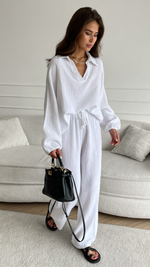 white textured co-ord set featuring a loose-fit collared blouse and drawstring wide-leg trousers, accessorized with a black leather handbag, layered gold necklaces, and black sandals