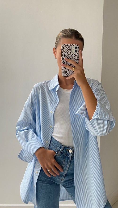 Woman wearing an oversized light blue and white striped button-down shirt over a white tank top, paired with high-waisted blue jeans.