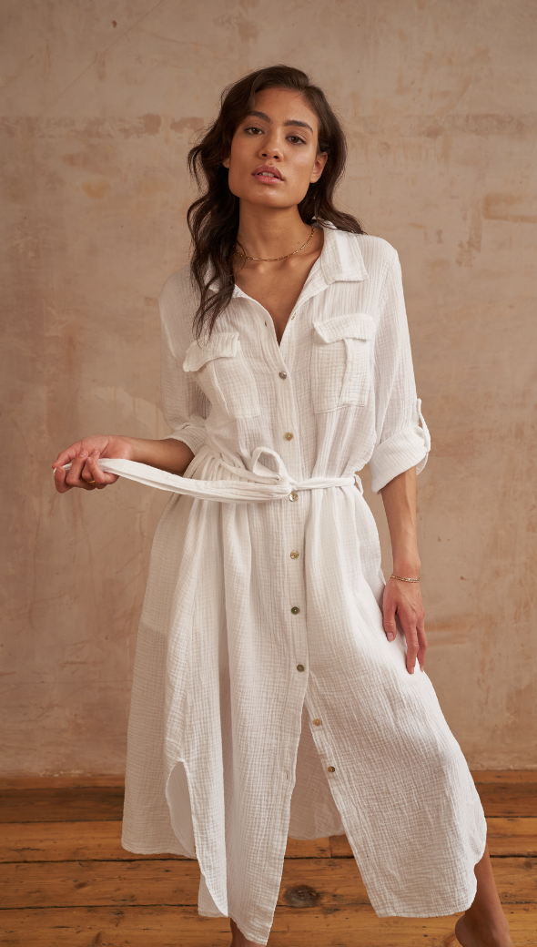 Woman wearing a white button-down shirt dress with a belted waist, rolled-up sleeves, and a textured gauze fabric