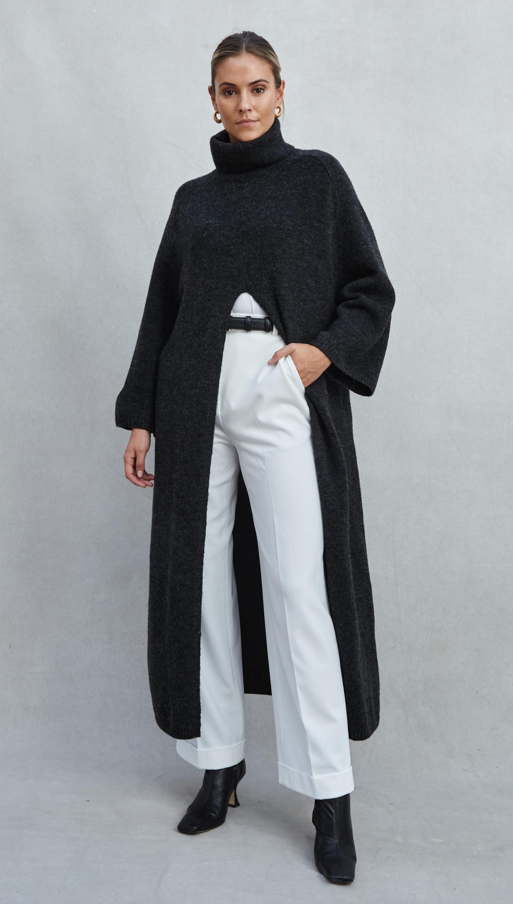 charcoal grey longline knit sweater with a turtleneck and front slit, styled with white tailored trousers, a black leather belt, and black ankle boots